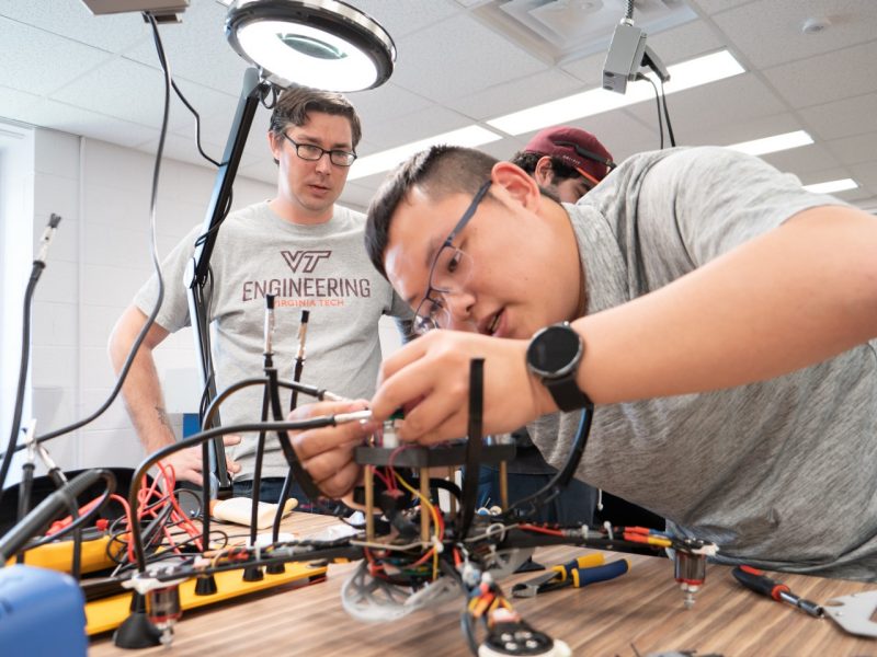 A student wiring together parts on a mechanical device while another student observes.