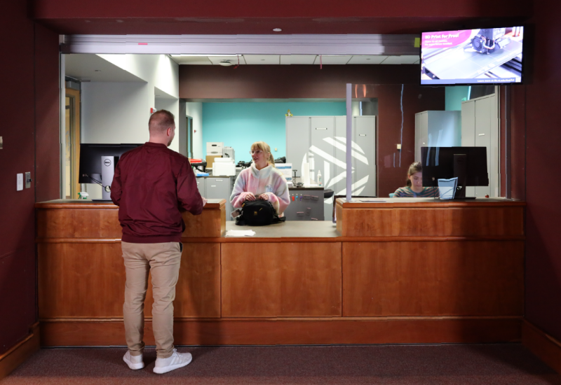 A patron stands at the Studios Technology Lending Desk, a smooth wood service point inset into maroon walls. Behind the desk are two student employees and many large aluminum cabinets.