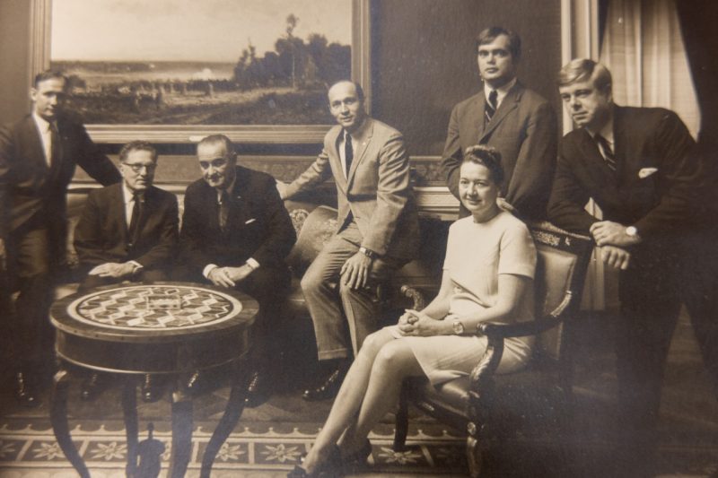 Genevieve Herrell sits in a chair surrounded by seated and standing men in suits including 26th President Lyndon B. Johnson.