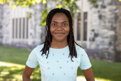 Imma Mwanja stands in front of Newman Library's long windows and Hokie Stone on a sunny day.