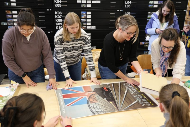 Four people stand on one side of a wooden table, touching and looking at a poster that is lying flat on the table and depicts a train.