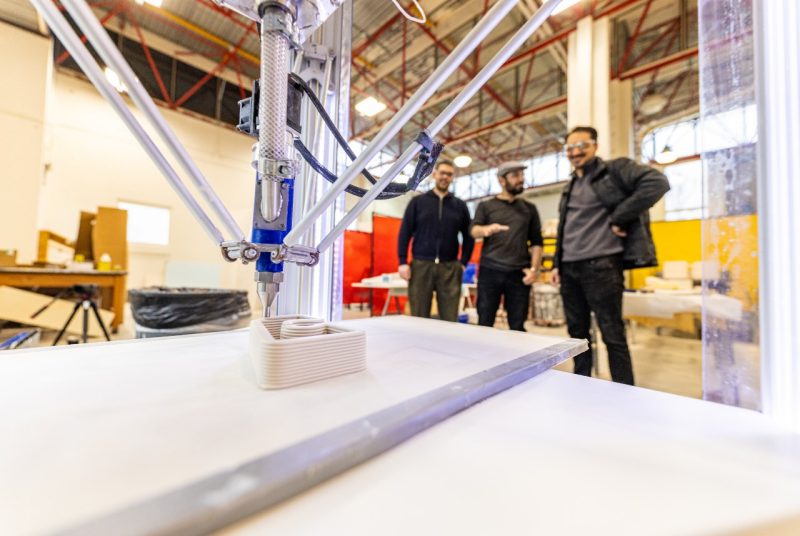 A close-up view of a 3D printer in action, extruding layers of material to form a rectangular object, with three researchers standing in the background observing the process in a brightly lit workshop.