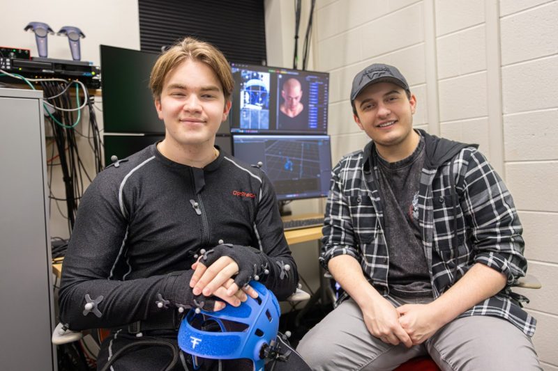 Interior of the Virtual Environments Studio where two young men are seated next to each other, smiling at the camera. On the left, a person is wearing a specialized black OptiTrack motion capture suit with numerous small gray spherical tracking markers attached to it at key points on the shoulders, arms, and body. He is holding a blue facial capture helmet device in his hands. On the right, a person is wearing a black and white plaid hoodie over a dark gray shirt and light gray pants. He is also wearing a black hat. Behind them is a workstation with multiple computer monitors displaying graphics related to their work. One screen shows a 3D facial model, while another shows a motion capture wireframe tracking points in a 3D space. The background features white tiled walls and various technical equipment, including virtual reality controllers in the upper left corner. Cables and computer hardware are visible on the left side of the image. 