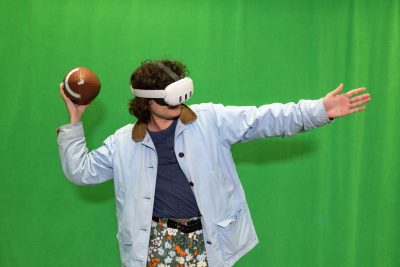 Student Michael  Davis-King wears virtual reality goggles and holds a football behind his shoulder in his right hand with his left arm straight in front of him acting out a football pass play in front of a green screen.