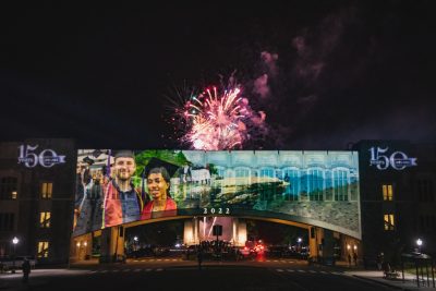 Images projected onto Torgersen Bridge while a firework explodes in the sky behind it. 