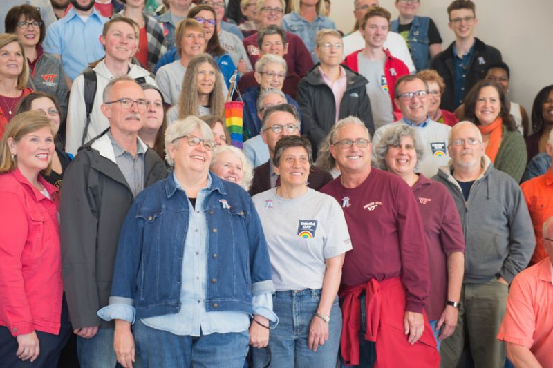 A smiling group gathered together for a photograph. Many of the members are wearing articles of denim, and shirts with a graphic that say "Denim Day at Virginia Tech" over a rainbow.