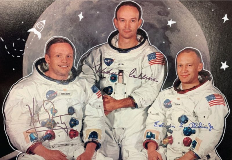 A signed photo of the Apollo 11 astronauts in their spacesuits, without helmets or gloves, sitting together smiling against a photo backdrop of the moon.