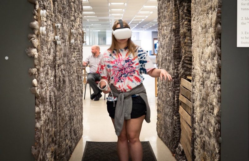 A young student wearing a virtual reality headset and holding a controller walks through the exhibit.