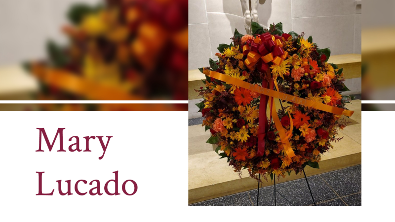 A large wreath including maroon and orange flowers and a maroon and orange bow placed on a stand.