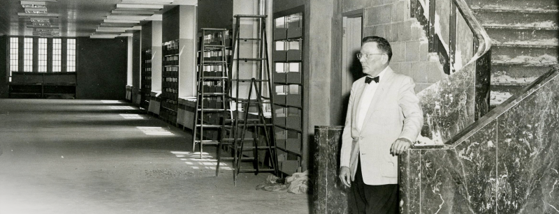 A man in a formal suit and bow tie stands at the bottom of a marble staircase overlooking a nearly completed, large, and empty library floor.