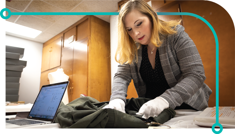 A researcher is leaning over an olive green jacket inspecting the garment using her white glove covered hands.