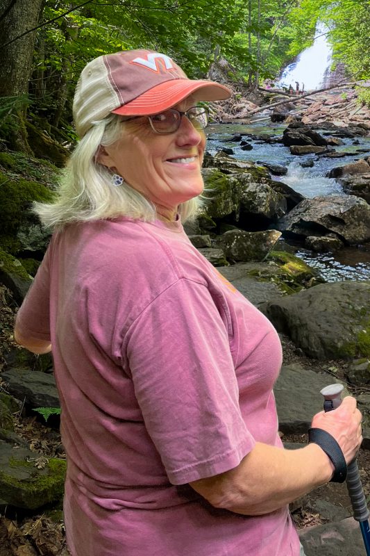 A woman wearing a Virginia Tech "VT" baseball cap looks over her shoulder to smile to the camera while hiking at the Cascades.