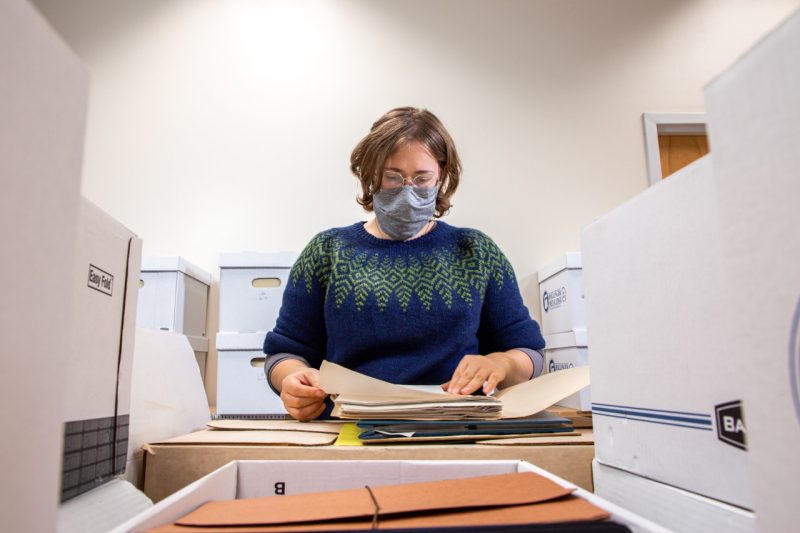 A masked woman stands between stacks of filing boxes and thumbs through a small stack of papers and folders placed on one stack.