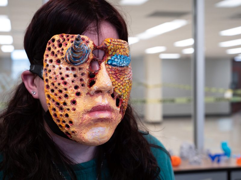 A student wearing a textured, painted mask depicting a person’s face which has several tiny holes throughout it.