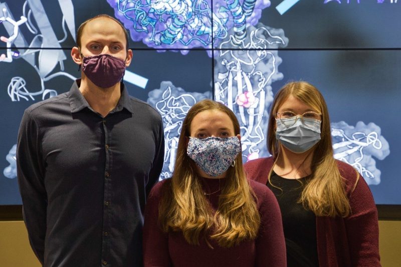 The three Virginia Tech authors, wearing masks, stand with their backs to a large video wall that is displaying a COVID-19 protein.