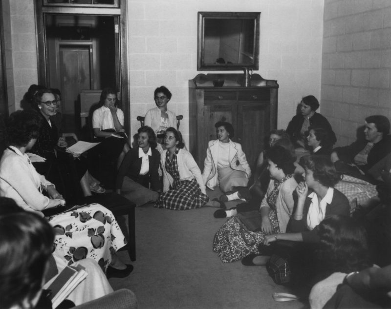 A group of women, some college students and some faculty, sitting in a room together smiling and talking.