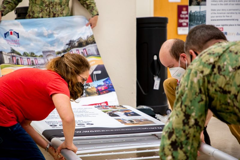 Four people, including two in military fatigues, crouched on the ground around a large metal frame, sliding the printed exhibit fabric onto the frame.