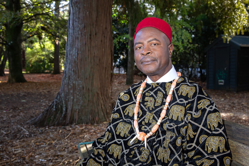 Agonzino wearing traditional Nigerian clothing sits on a wood bench in front of a wooded area.