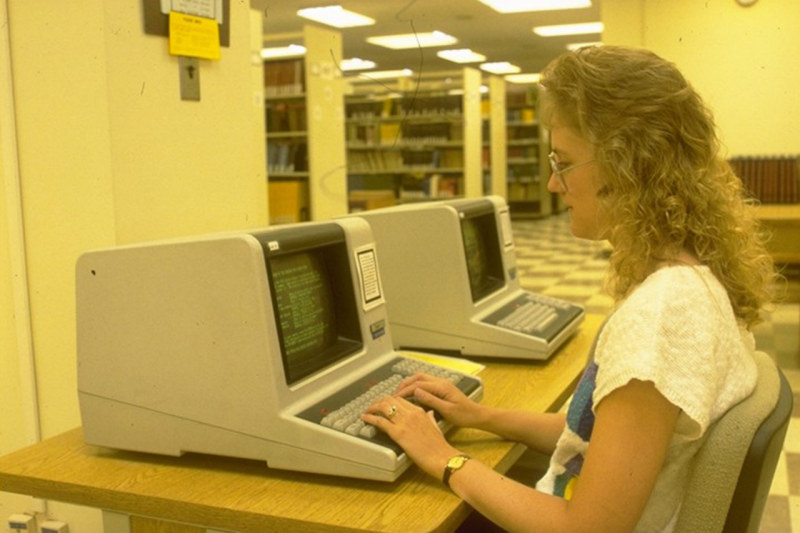 A person typing alone on a compact CRT all-in-one computer located at the end of a row of bookshelves.