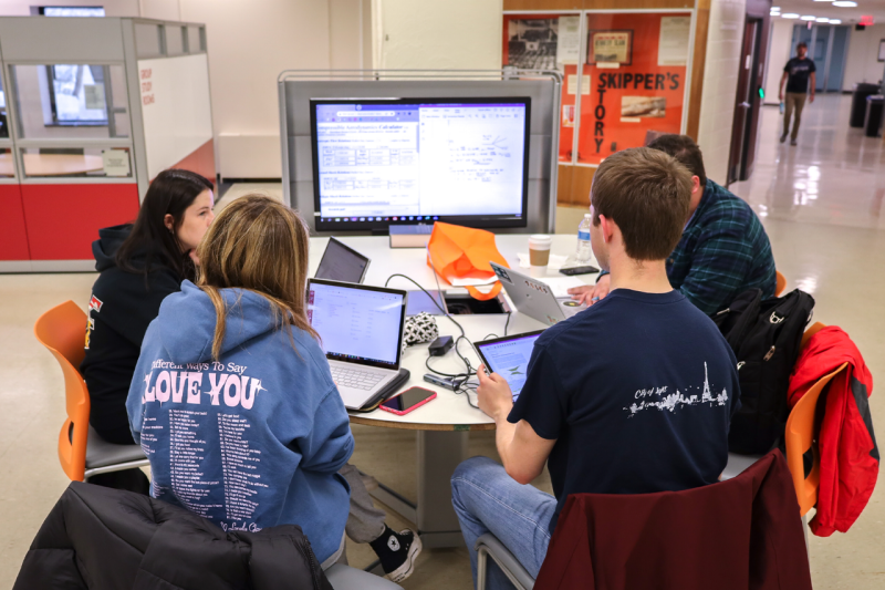 A group of students working together on their laptops at a media table with a large monitor.