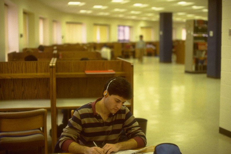 A person wearing headphones writing on paper alone on the 3rd floor of Newman Library. They are sitting at one of several wooden study carrels arranged around a curved wall.