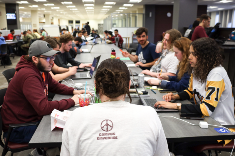 A large group of students sitting together at a long table talking and working on their laptops. More large groups of students are spread out at scattered tables with power outlets.