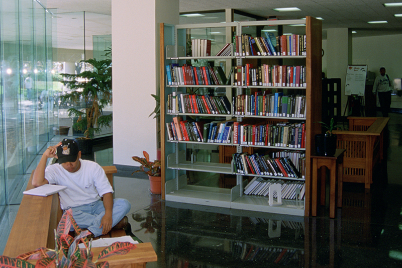 A person sitting sideways on a cushioned bench against a glass wall reading from their notebook. Next to them in the space are a few bookshelves and potted plants.