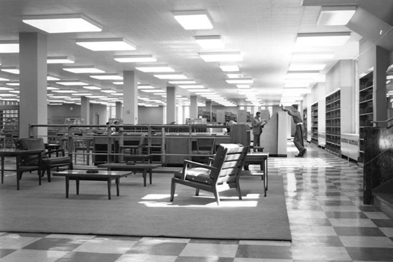 An open, tiled, indoor space with a few chairs and tables on sections of carpet. Two cadets lean against a short wall in the distance and bookshelves line the walls.