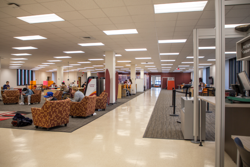 An open, tiled and carpeted space with many students at clusters of chairs and tables. A print center, laptop kiosks, and media surround the furniture and line the walls.