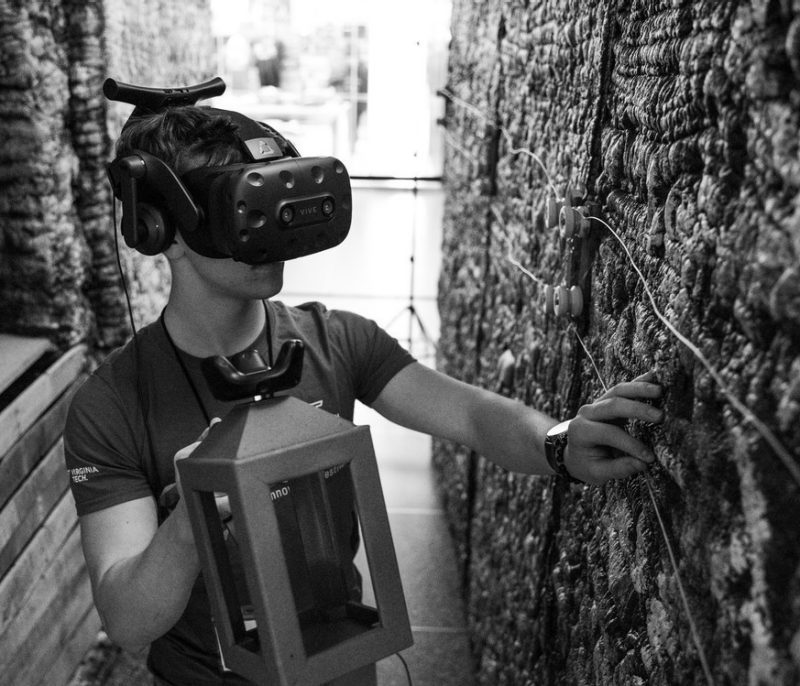 Photo of student holding a VR remote while wearing a headset. Student is inside of a replica of a war tunnel that they can feel as they see it in VR.